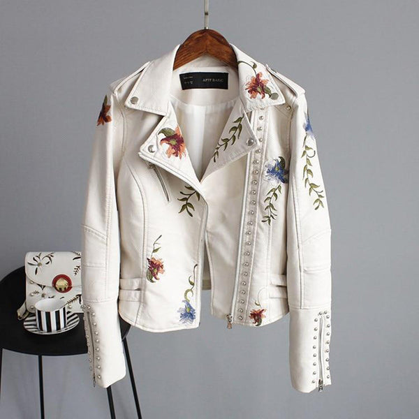 Beige leren jas met bloemenborduursels, zilveren studs en asymmetrische rits, hangend aan een houten hanger tegen een grijze achtergrond.