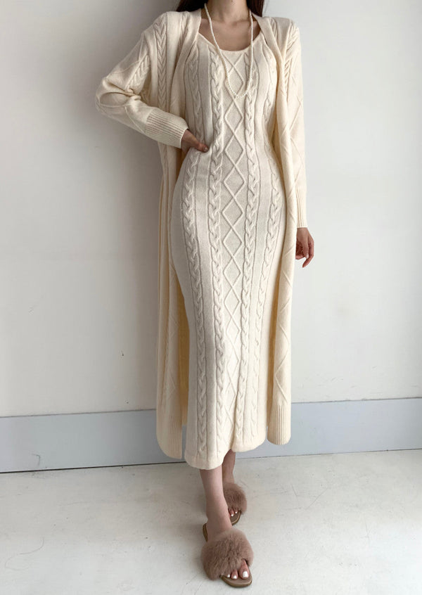 Vrouw in beige gebreide jurk met kabelpatroon en lange mouwen, gecombineerd met bijpassende lange vest en pluizige slippers, tegen witte achtergrond.