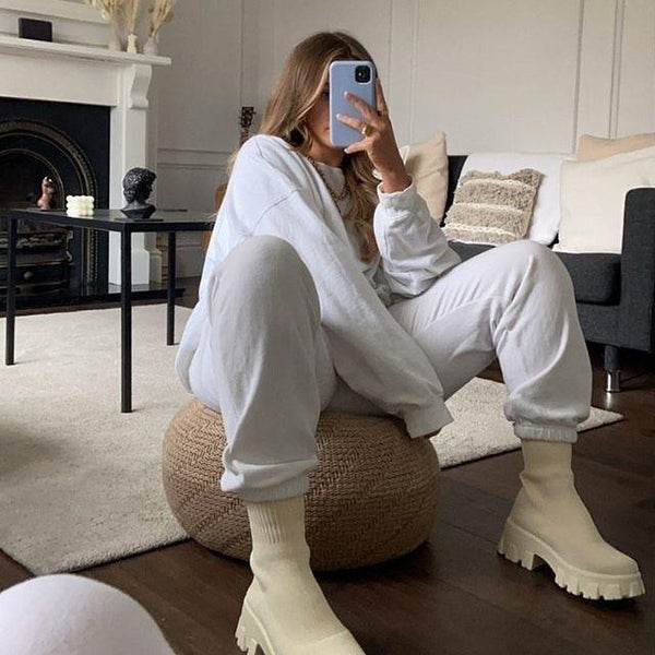 Vrouw in witte loungewear en beige laarzen, zittend op een poef in een moderne woonkamer. Trendy mode, comfortabele kleding, minimalistisch interieur.