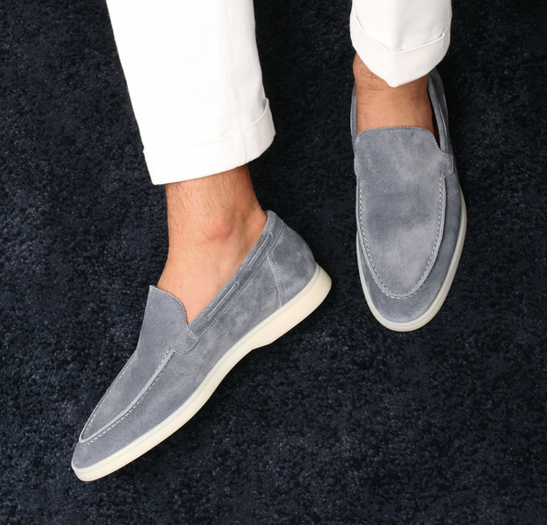 Grijze suède loafers voor heren op donkerblauw tapijt, gedragen met witte broek. Stijlvolle casual schoenen, comfortabele pasvorm.