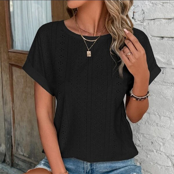 Vrouw draagt zwarte kanten blouse met korte mouwen, gouden ketting en armband, casual zomeroutfit, tegen witte bakstenen muur.