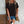 Vrouw in zwarte T-shirt en denim shorts, met bruine schoudertas en koffiehouder, tegen witte bakstenen muur. Casual zomeroutfit, modeaccessoires.