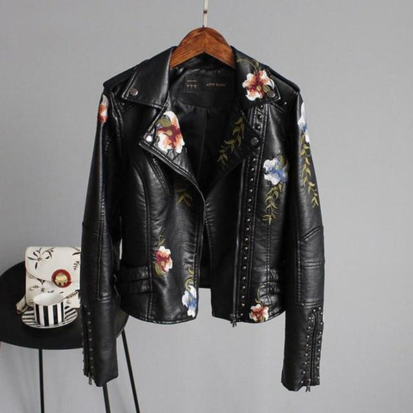 Zwarte leren jas met bloemenborduursels, studs en asymmetrische rits, hangend aan een houten hanger tegen een grijze achtergrond.