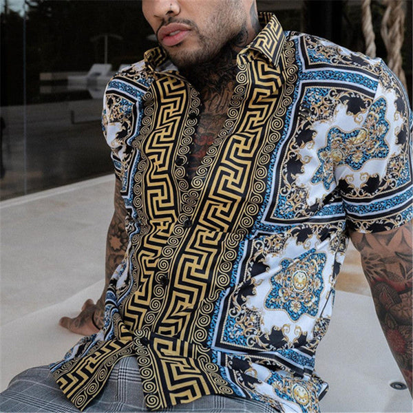 Man in kleurrijke, barokke print blouse met blauwe en gouden patronen, zittend op een moderne achtergrond. Stijlvolle herenmode, zomertrends.