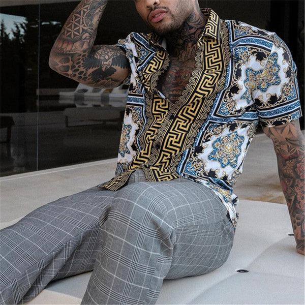 Man met getatoeëerde armen draagt een blauw-witte blouse met gouden patronen en grijze geruite broek, zittend op een moderne bank.