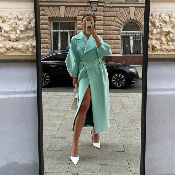 Vrouw in mintgroene trenchcoat met split, witte hakken en tas, poseert voor spiegel op straat. Stijlvolle mode, stedelijke achtergrond.