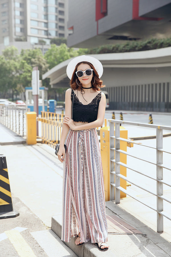 Vrouw in boho-broek met streepjespatroon, zwarte crop top en witte hoed, wandelend in stedelijke omgeving. Zomerse mode, casual stijl.