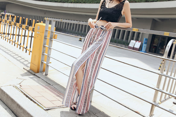 Vrouw in boho-broek met kleurrijk patroon en hoge split, draagt een zwarte kanten crop top, leunt tegen een metalen hek in een stedelijke omgeving.