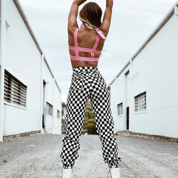 Vrouw in roze top en zwart-wit geruite broek poseert op industriële locatie. Modieuze streetwear, trendy outfit, urban stijl.