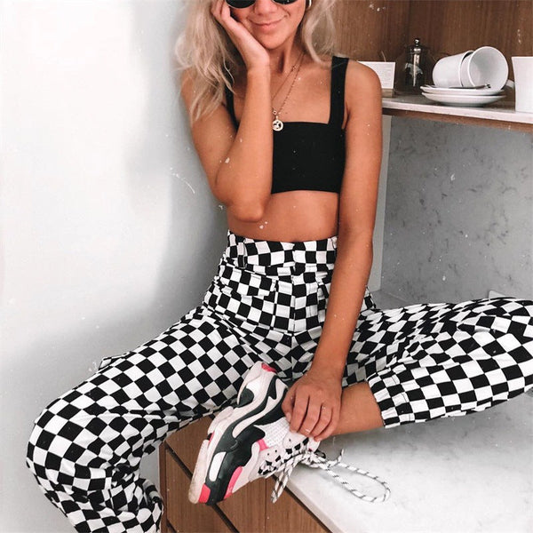 Vrouw in zwart crop top en geruite broek, zittend op keukenblad. Modieuze outfit, casual stijl, trendy sneakers. Perfect voor streetwear inspiratie.