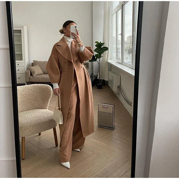 Vrouw in stijlvolle bruine trenchcoat en wijde broek, poseert voor spiegel in modern interieur. Trendy mode, herfstkleding, minimalistisch design.