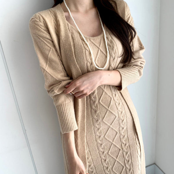 Vrouw in beige gebreide jurk met kabelpatroon en lange mouwen, draagt een parelketting. Stijlvolle herfstmode, elegant en trendy design.