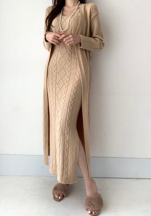 Vrouw in beige gebreide jurk met kabelpatroon en lange mouwen, gecombineerd met bijpassende lange vest en pluizige slippers. Elegante herfstmode.