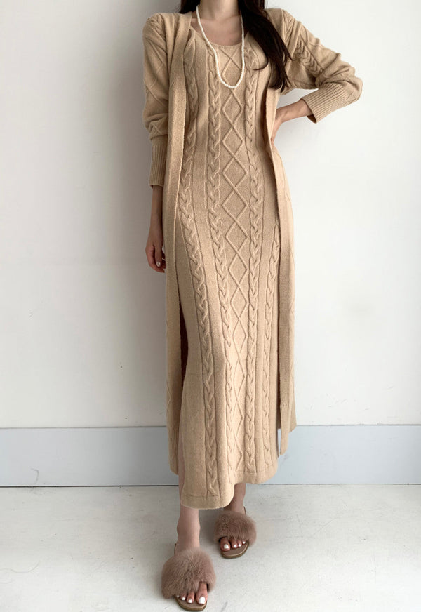 Vrouw in beige gebreide maxi-jurk met kabelpatroon, lange mouwen en split, gecombineerd met pluizige slippers. Stijlvolle herfstmode.