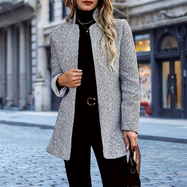 Vrouw in stijlvolle grijze visgraatjas, zwarte coltrui en broek, met handtas, op stedelijke achtergrond. Mode, herfstkleding, elegante damesjas.