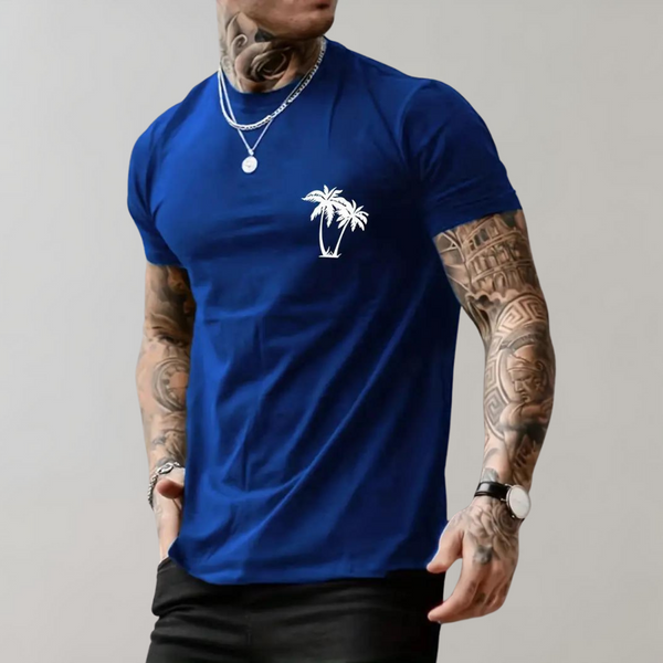 Man in koningsblauw T-shirt met palmboomprint, tatoeages, zilveren kettingen en horloge. Casual mode, herenkleding, trendy stijl.