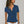 Vrouw in blauwe blouse met korte mouwen, knoopsluiting en borstzakken, draagt witte broek en lange ketting. Casual damesmode, zomeroutfit.