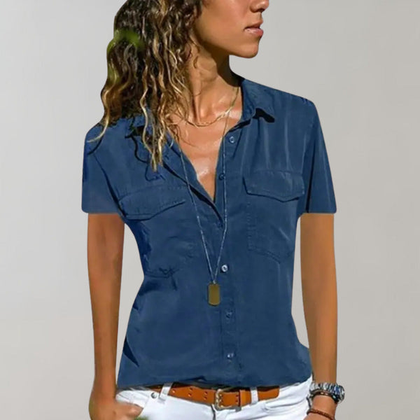 Vrouw in blauwe blouse met korte mouwen, knoopsluiting en borstzakken, draagt witte broek en lange ketting. Casual damesmode, zomeroutfit.