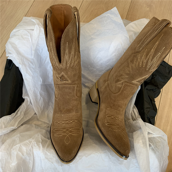 Bruine suède dameslaarzen met westernstijl, middelhoge hak en sierstiksels, verpakt in wit papier op houten vloer. Perfect voor herfstmode.