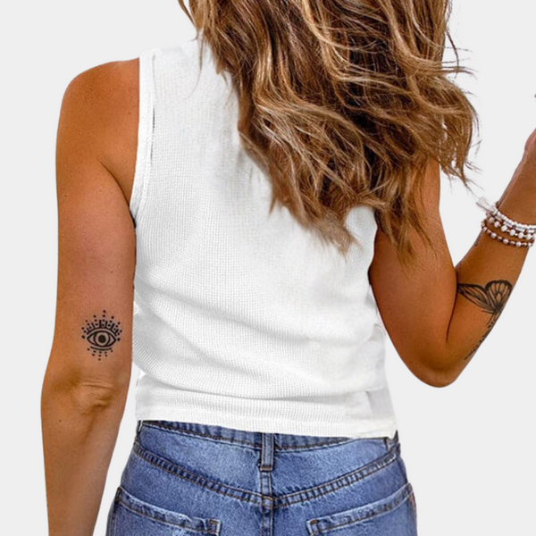 Vrouw in witte tanktop en blauwe jeans, met golvend haar en armbanden, toont rug en tatoeage van oog op arm. Casual mode, zomeroutfit.