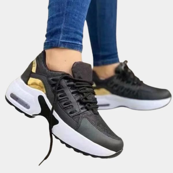 Zwarte sneakers met witte zool en gouden details, gedragen door persoon in blauwe jeans. Stijlvolle sportschoenen voor dagelijks gebruik.