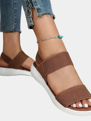 Bruine sandalen met elastische banden en witte zool, gedragen door een persoon met enkelbandje en jeans. Comfortabele zomerschoenen voor dames.