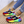 Kleurrijke sneakers met regenboogzool, blauwe en rode accenten, gedragen door persoon met opgerolde jeans en gouden enkelbandjes op houten vloer.