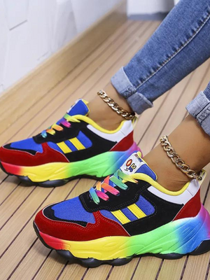 Kleurrijke sneakers met regenboogzool, blauwe en rode accenten, gedragen door persoon met opgerolde jeans en gouden enkelbandjes op houten vloer.