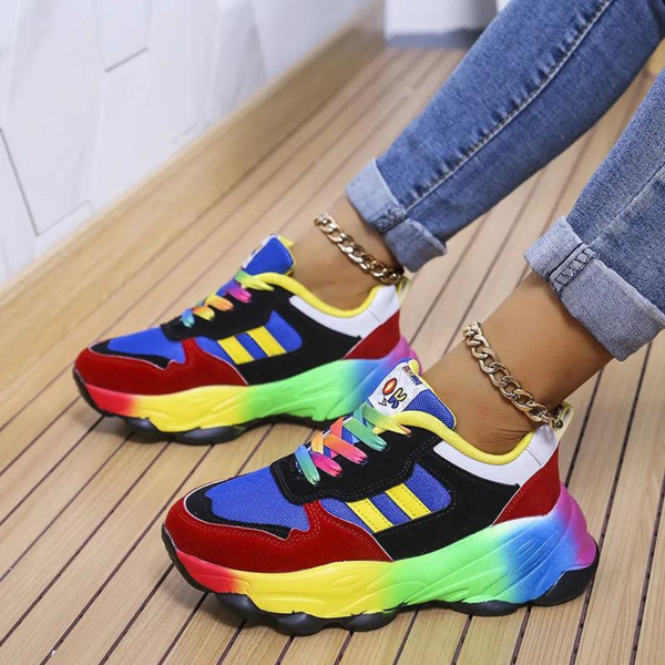 Kleurrijke sneakers met regenboogzool, blauwe en rode accenten, gedragen door persoon met opgerolde jeans en gouden enkelbandjes op houten vloer.