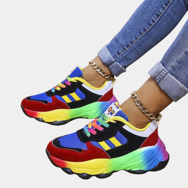 Dames sneakers met regenboogkleuren, blauwe en rode accenten, gele strepen, gedragen met opgerolde jeans en gouden enkelbandjes.