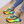 Groene sneakers met regenboogzool, kleurrijke veters en gouden enkelbanden, gedragen door persoon in opgerolde jeans op houten vloer.