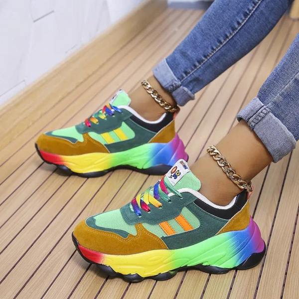 Groene sneakers met regenboogzool, kleurrijke veters en gouden enkelbanden, gedragen door persoon in opgerolde jeans op houten vloer.