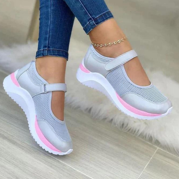Vrouw draagt grijze en roze sportieve Mary Jane schoenen met klittenband, gecombineerd met blauwe jeans en enkelbandje, op houten vloer.