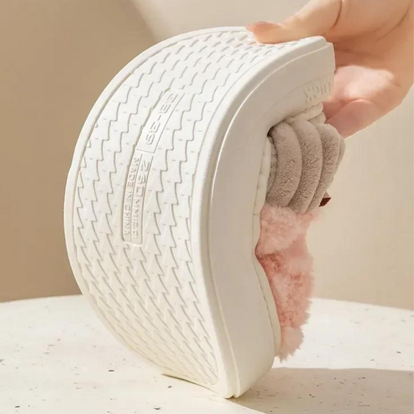 Hand houdt gebogen witte slipper met zachte roze en grijze pluche voering vast, op een lichte achtergrond. Comfortabele damespantoffel, antislip zool.