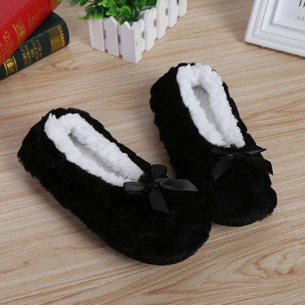 Zwarte pluche pantoffels met witte voering en strikje, op houten vloer. Comfortabele dameshuisschoenen, perfect voor winterse warmte.