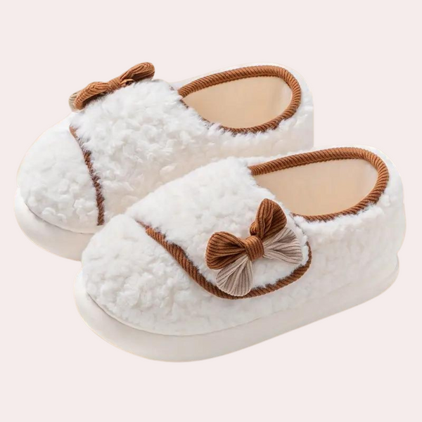 Witte pluche babyslofjes met bruine strikjes, warme voering en antislipzool. Perfect voor comfort en stijl, ideaal als kraamcadeau.