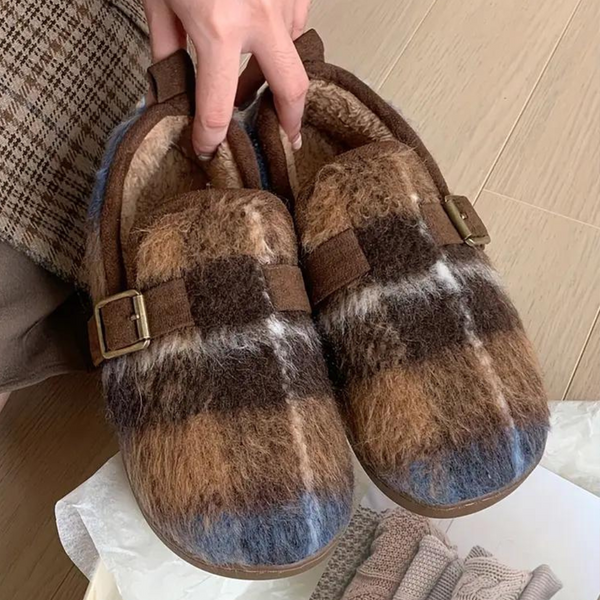 Hand houdt bruine en blauwe wollen pantoffels met gesp vast op houten vloer. Comfortabele, warme huisschoenen voor winterse dagen.