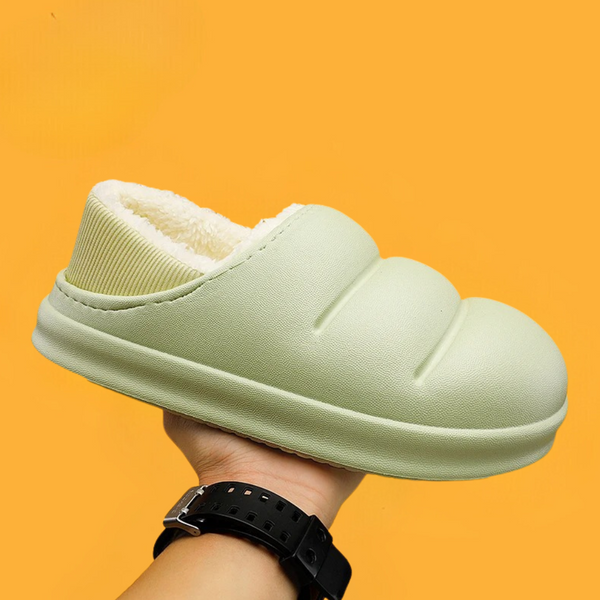 Lichtgroene pluche pantoffel met gewatteerd ontwerp, warme voering en antislipzool, vastgehouden door een hand tegen een oranje achtergrond.