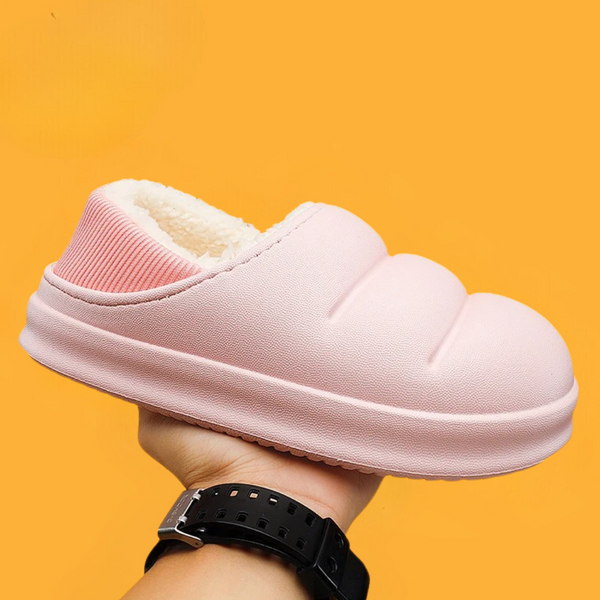 Roze pluche pantoffel met gewatteerd ontwerp, warme voering en rubberen zool, vastgehouden tegen een oranje achtergrond. Comfortabele huispantoffel.
