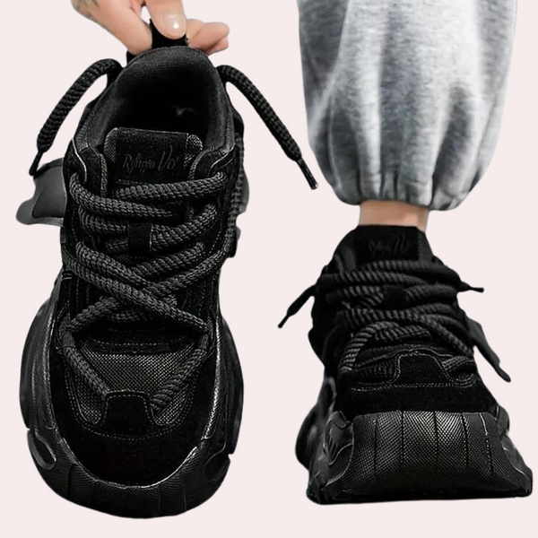 Zwarte chunky sneakers met dikke zool en veters, gedragen door een persoon in grijze joggingbroek. Trendy sportschoenen voor dagelijks gebruik.