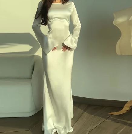 Vrouw in elegante, lange witte satijnen jurk met wijde mouwen, staand in moderne woonkamer. Perfect voor avondkleding en formele evenementen.