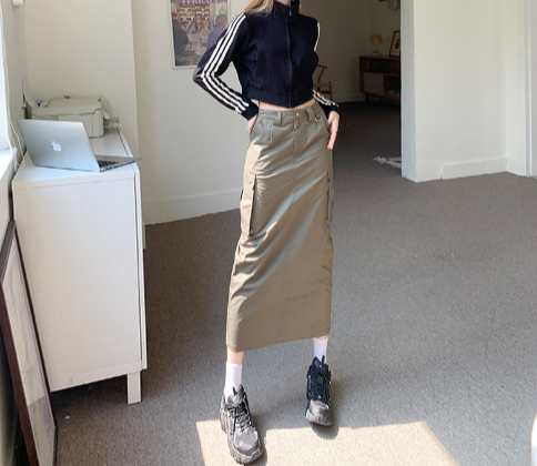 Vrouw draagt lange cargorok met zakken, gecombineerd met zwart sportjack en sneakers. Modieuze outfit in moderne kamer.