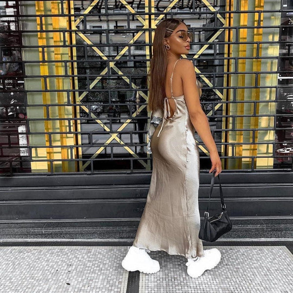 Vrouw in champagnekleurige jurk met open rug, witte sneakers en zwarte handtas voor een moderne architecturale achtergrond. Fashion, street style.