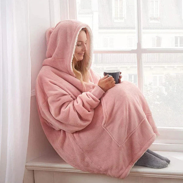 Vrouw in roze oversized hoodie deken met capuchon, zittend op vensterbank, genietend van warme drank. Comfortabele loungewear, knusse winterstijl.