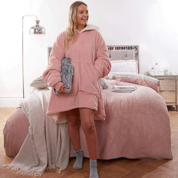 Vrouw in roze oversized hoodie deken staat in slaapkamer, naast bed met roze dekbedovertrek. Comfortabele, warme kleding voor thuis.