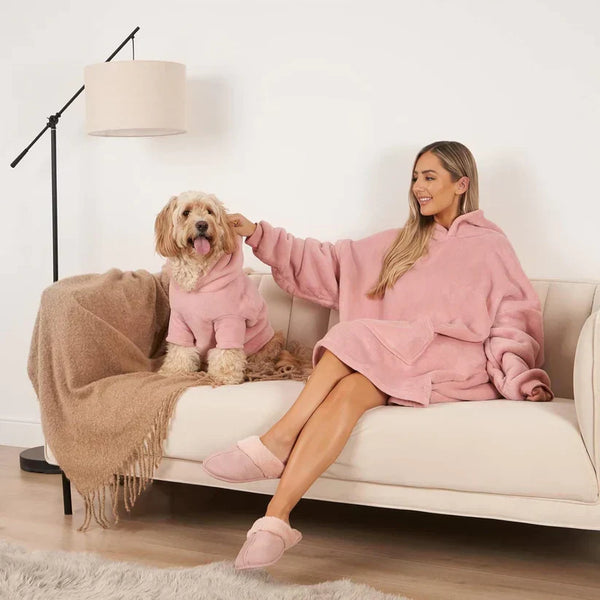 Vrouw en hond in roze fleece hoodies op bank, moderne woonkamer, vloerlamp, beige deken, comfortabele slippers, huiselijke sfeer.