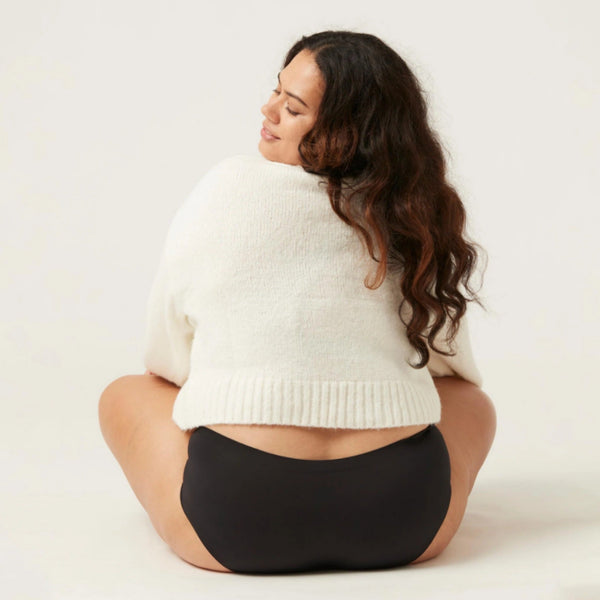 Vrouw in witte trui en zwarte slip, zittend op de grond. Comfortabele damesmode, zachte materialen, trendy kledingstijl.