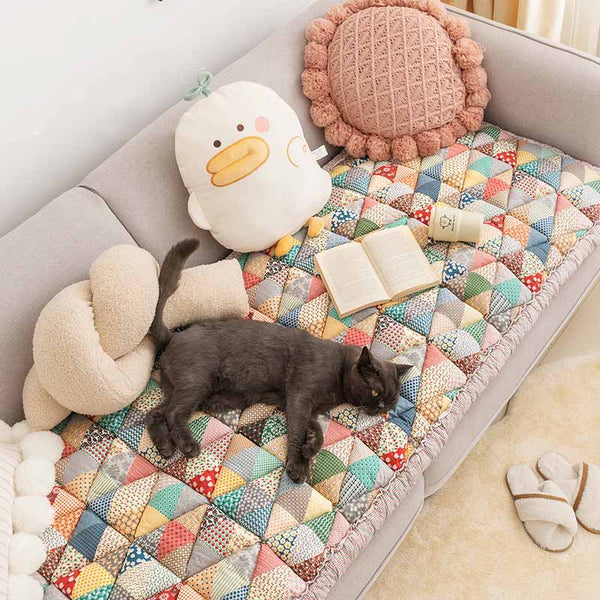 Grijze kat op kleurrijke katoenen bankhoes met patchwork patroon, naast een open boek en pluche speelgoed. Perfecte bankbescherming voor huisdieren.