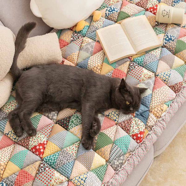 Grijze kat op kleurrijke patchwork bankhoes van katoen, naast open boek en knuffelbeer. Perfecte bankbescherming voor huisdieren.