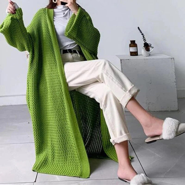 Vrouw in groene gebreide lange cardigan, witte broek en slippers met bont, zittend in moderne woonkamer. Trendy damesmode, comfortabele stijl.